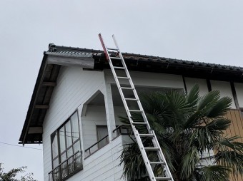 二階の瓦屋根の軒先に二連梯子を掛けた様子